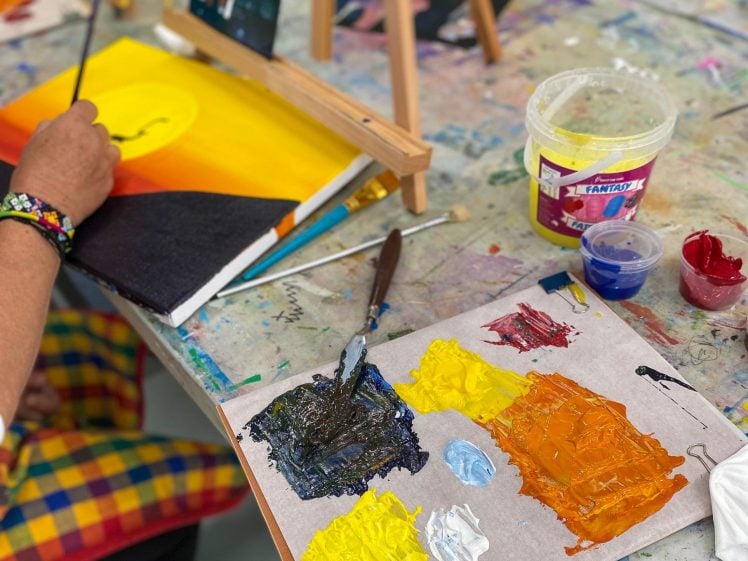 Image of A person in colourful chequered trousers paints a sunset scene on a canvas, with paintbrushes, a palette of mixed colours, and paint containers on a messy art table.