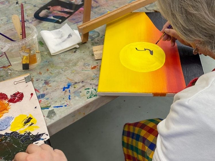 Image of A person with short grey hair paints a yellow circle with a black shape inside on a canvas with a red, orange, and black background. Art supplies, brushes, and a palette with various paint colours are on the table.