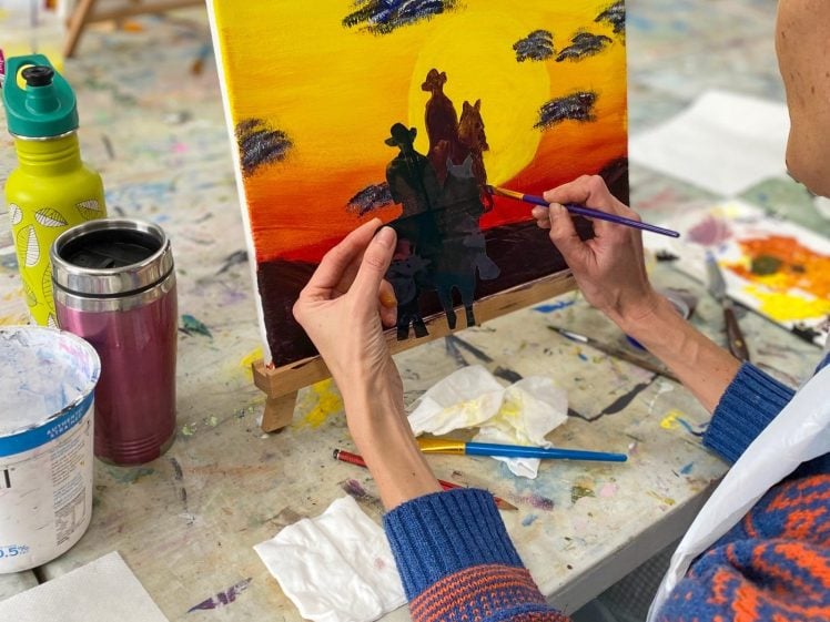 Image of A person in a blue and orange jumper paints a sunset scene with silhouettes of people and trees on a canvas at an art class. Art materials, brushes, and cups are scattered on the table.