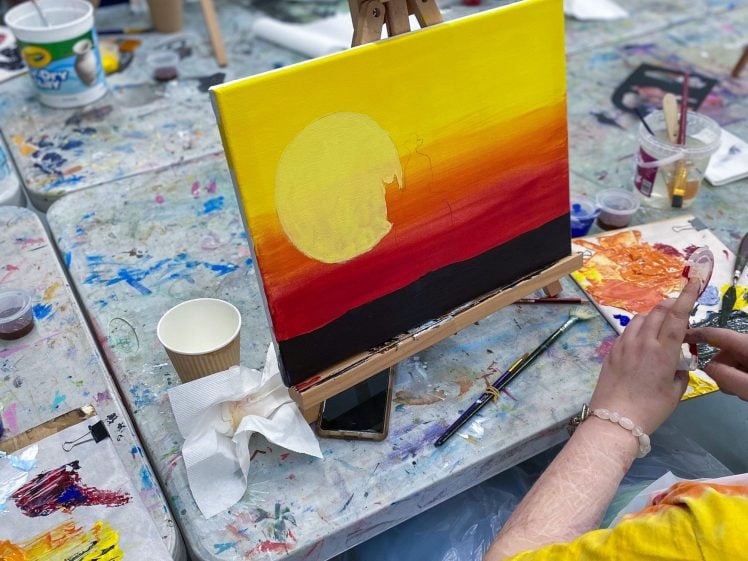 Image of A person paints a vivid sunset scene on canvas, with bright yellow, orange, and red colours. The painting is on an easel, surrounded by art supplies, brushes, and paint palettes on a splattered, messy table.