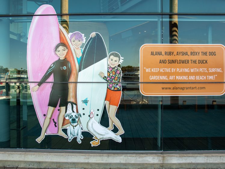 Image of A window displays a mural of three children with surfboards, a dog, and a duck. Next to them is a sign with their names and a quote about staying active through surfing, pets, gardening, art, and beach time.