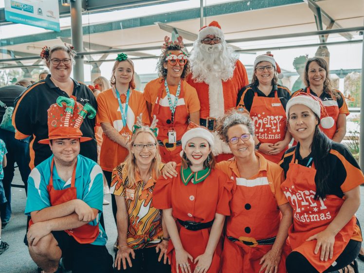 Image of A group of people dressed in festive Christmas attire, including Father Christmas, elves, and shirts with holiday designs, pose and smile for a photo indoors. Some wear hats, headbands, and cheerful accessories.