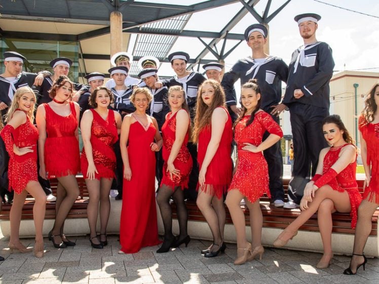 Image of A group of women in red dresses and men in sailor uniforms pose together outdoors, smiling at the camera. The women stand or sit in front, whilst the men stand behind them under a metal canopy.
