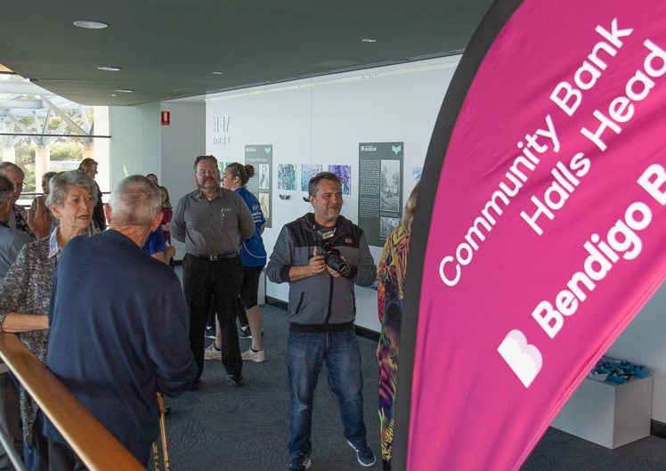 Image of A group of people gathers indoors near a large pink flag displaying Community Bank Halls Head Bendigo Bank. Some people are talking, while others look at displays on the wall.