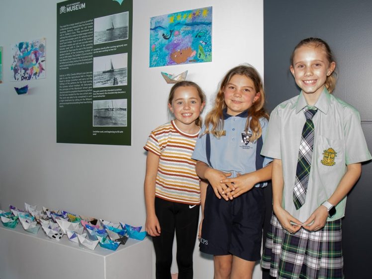 Image of Three smiling children stand indoors next to a display of colourful paper boats and artwork on a wall, with an information panel in the background. Two wear casual clothes; one wears a school uniform.