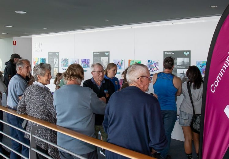 Image of A group of people stand in a queue inside a modern building, viewing artwork on a white wall. In the foreground is a pink banner with “Community Bank Halls Head Bendigo Bank” written on it.