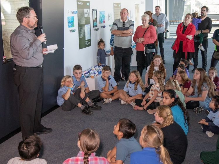 Image of A man speaks into a microphone whilst holding papers, addressing a seated group of children and standing adults in an indoor educational setting with posters on the wall behind him.