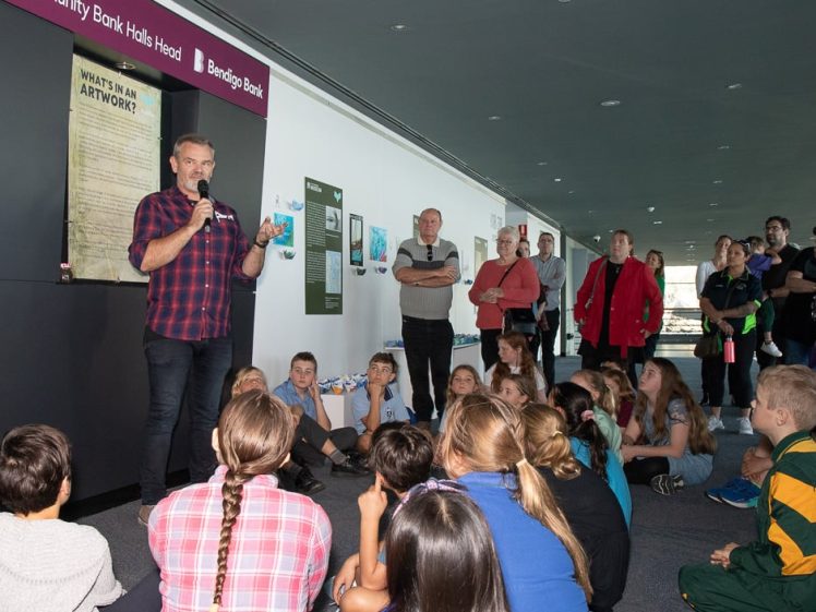 Image of A man speaks into a microphone in front of an audience of children seated on the floor, whilst adults stand and watch in the background inside a modern building. Posters are displayed on the wall behind him.