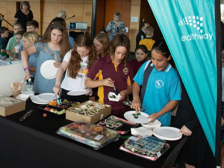 Image of A group of students stands at a buffet table, serving themselves food onto plates at an event. Packaged sushi and trays of appetisers are visible, with a turquoise Healthway banner in the foreground.
