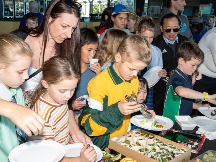 Image of A group of children and adults gather around a table, selecting food from a variety of snacks and sushi rolls at a busy indoor event. The atmosphere appears lively and sociable.