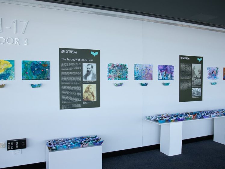 Image of A museum exhibit wall displays panels with text and historical photos titled The Tragedy of Black Bess, colourful artwork, and shelves filled with numerous paper cranes.