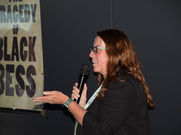 Image of A woman with long brown hair and glasses speaks into a microphone, gesturing with her hand. Beside her is a poster that reads THE TRAGEDY OF BLACK BESS against a dark background.