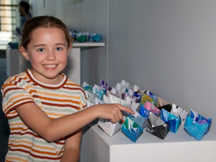 Image of A smiling girl in a striped shirt points to a colourful display of origami paper boats arranged on white plinths in an indoor setting.