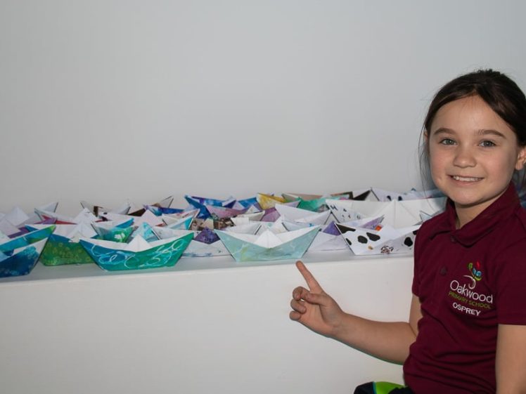 Image of A young girl in a maroon Oakwood Osprey shirt smiles and points to a row of colourful paper boats displayed on a white ledge against a plain wall.
