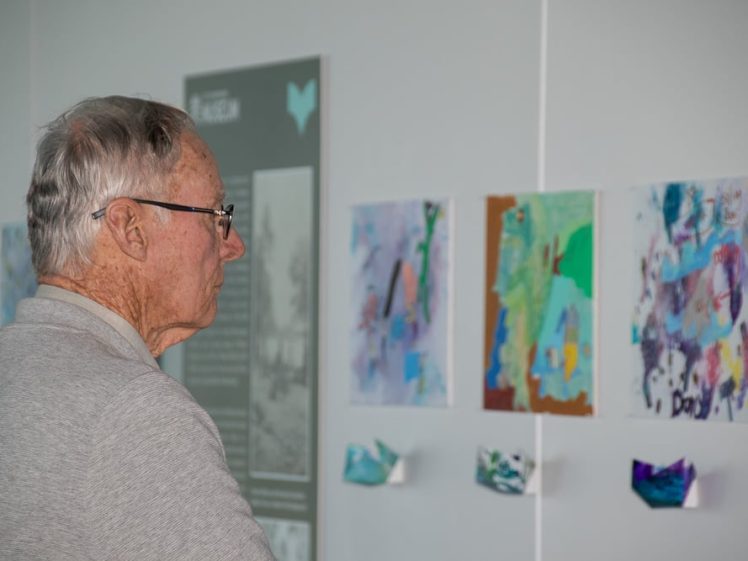 Image of An elderly man with grey hair and spectacles observes colourful abstract artwork displayed on a white wall in a gallery.