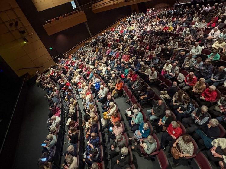 Image of A large audience sits in rows of seats inside a theatre, facing the stage. The venue is well-lit, and most seats are occupied by people of various ages, some using phones or talking quietly.