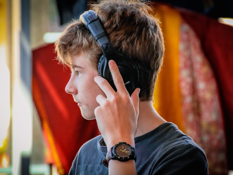 Image of A young person with light brown hair wearing a black T-shirt and headphones listens intently, touching the headphones with one hand. The background is softly blurred with warm colours.