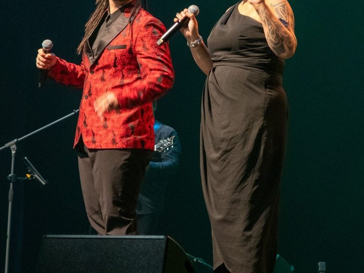 Image of A man in a red patterned jacket and a woman in a black dress sing on stage, each holding a microphone, with stage lights illuminating them and musical equipment visible in the background.