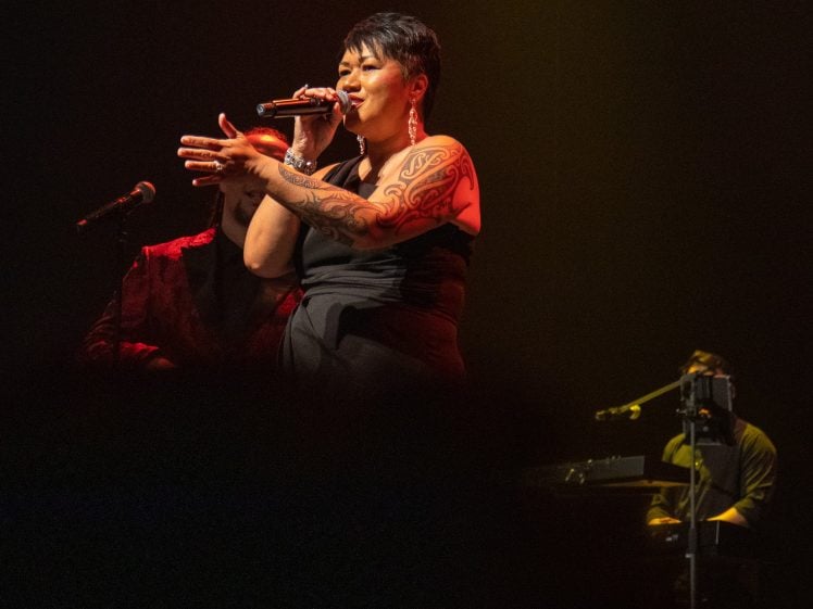 Image of A woman with short hair and a tattooed arm passionately sings into a microphone on stage, wearing a black outfit. She is illuminated by stage lights, with a keyboard player visible in the background.