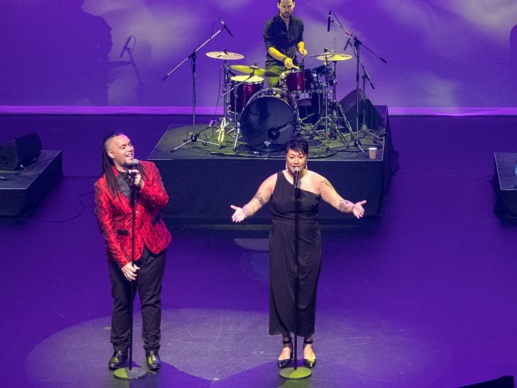 Image of Two singers perform on stage under purple lighting, one in a red jacket and black trousers, the other in a black dress, whilst a drummer plays in the background. They are illuminated by spotlights.