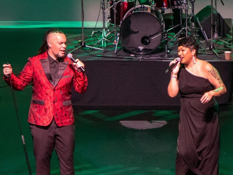 Image of Two singers perform on stage with microphones. The man wears a red patterned suit jacket and black trousers; the woman wears a black dress with a tattoo visible on her arm. Drums and stage lights are visible in the background.