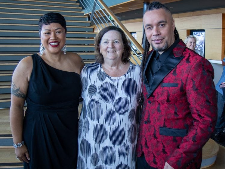 Image of Three people pose and smile together indoors near a staircase. The person on the left wears a black dress, the middle person wears a white dress with black spots, and the right person wears a red and black patterned suit.