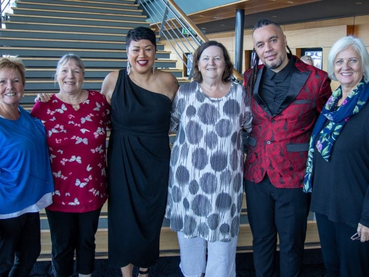 Image of Six adults stand smiling together indoors in front of a wooden staircase. They are dressed in a mix of colourful and patterned clothing, and the setting appears modern and bright.