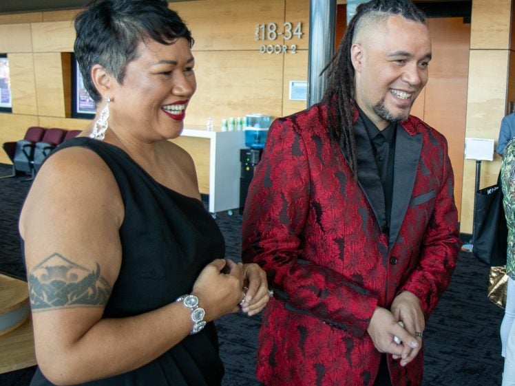 Image of Two people dressed formally are smiling and laughing indoors. The woman wears a black dress, earrings, and has a tattoo on her upper arm. The man wears a red patterned blazer and has long plaits. They stand near a wooden wall with door signs.