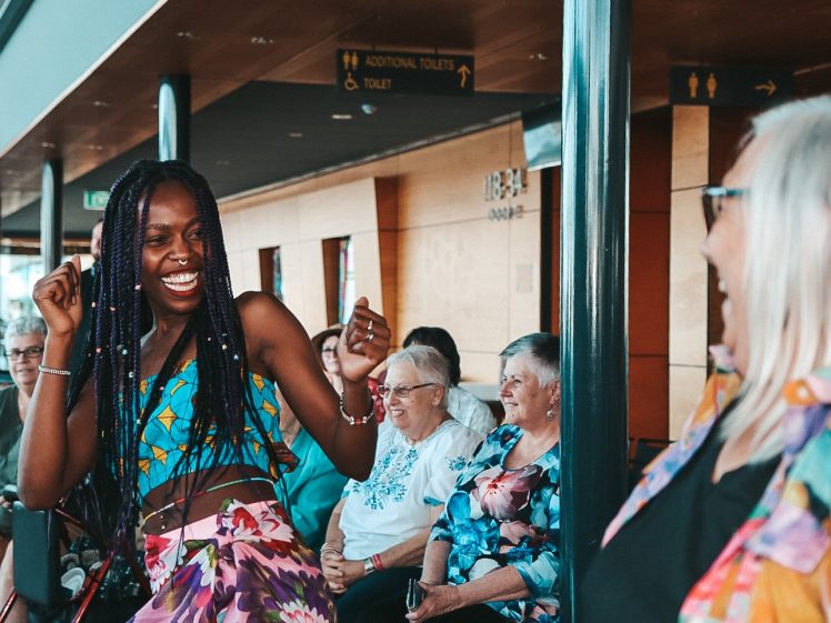 Image of A woman in colourful clothing joyfully dances in front of a smiling group of seated elderly women, who are watching her performance indoors. Another person in the foreground is also smiling.