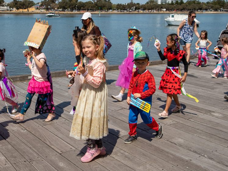 Image of Children dressed in colourful costumes, including princesses, superheroes, and fairies, walk in a parade on a wooden boardwalk by a river on a sunny day. Adults accompany them in the background.