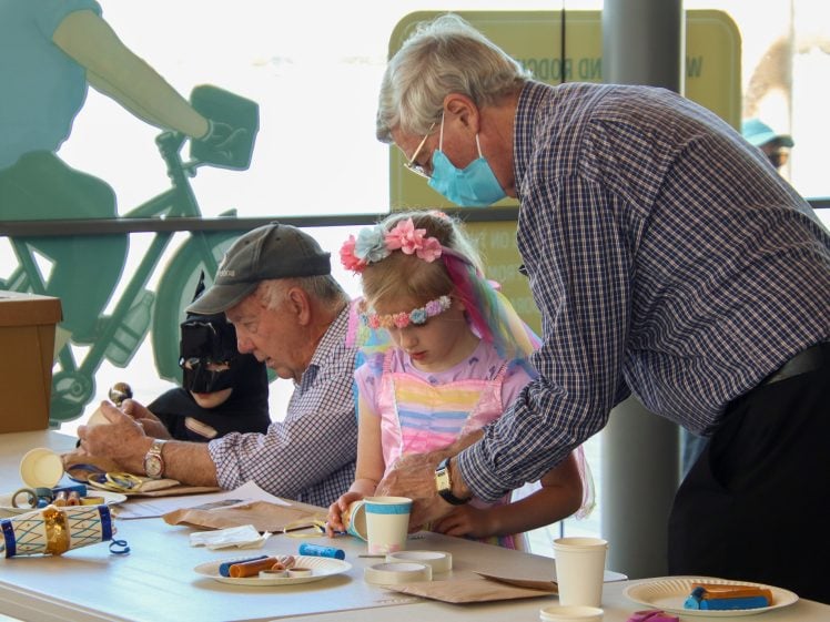 Image of An older man helps a young girl wearing a flower headband with a craft project at a table, whilst another man works beside them. Art supplies and finished crafts are on the table.
