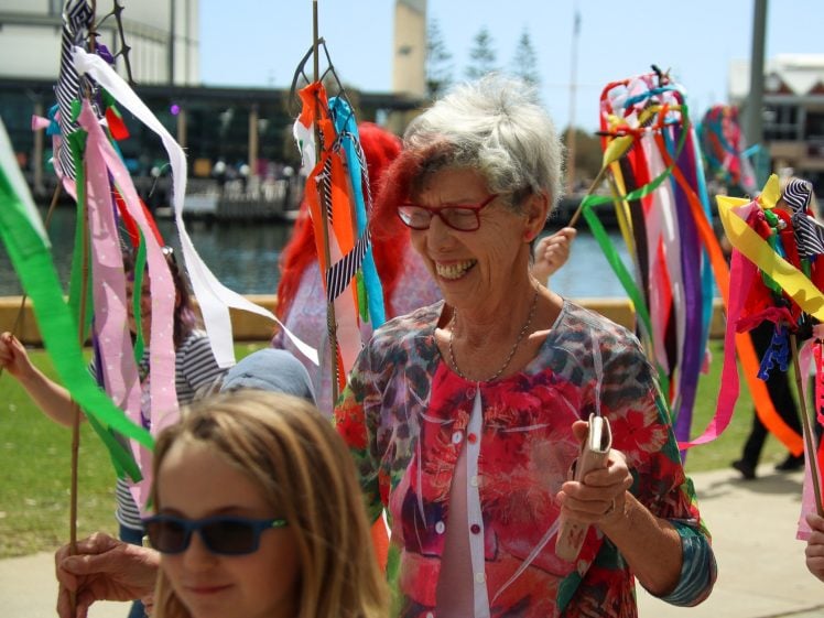 Image of An older woman with glasses, smiling, walks outdoors holding colourful ribbon streamers. Children around her also carry streamers. It appears to be a festive, sunny day near a waterfront.