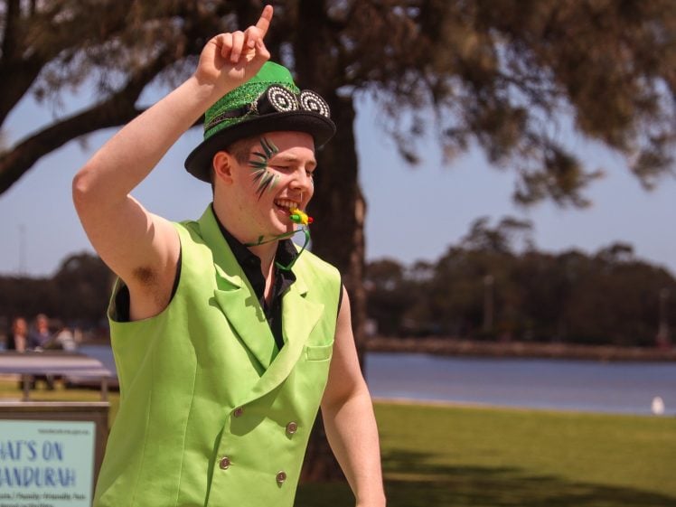Image of A performer wearing a bright green waistcoat and hat with painted face markings gestures enthusiastically outdoors, holding a whistle in their mouth, with trees and water in the background.