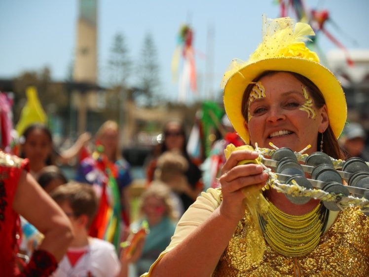 Image of A woman in a bright yellow hat and costume smiles whilst shaking a percussion instrument during a colourful outdoor festival, with other people and festive decorations in the background.