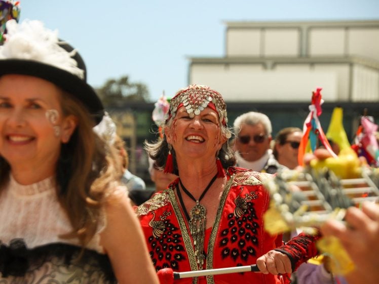Image of A group of people in colourful, festive costumes smile and parade outdoors. The focus is on a woman in red with a jewelled headdress, holding a musical instrument, surrounded by cheering participants.