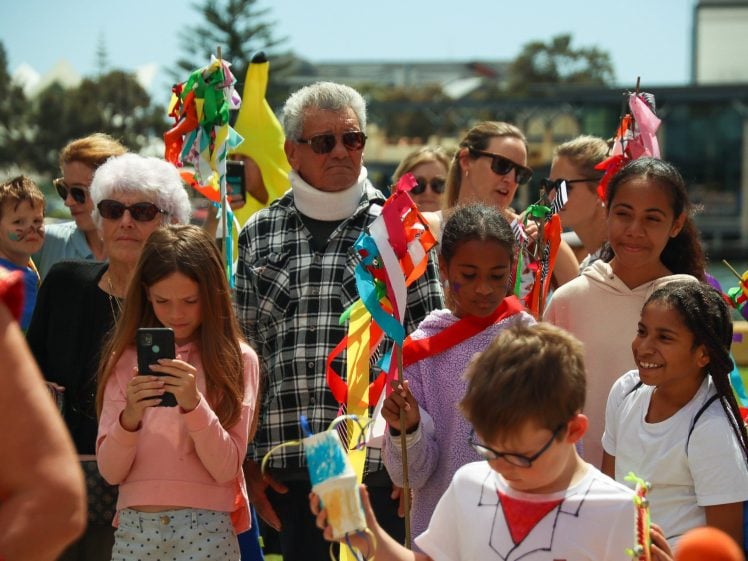 Image of A diverse group of children and adults gathers outdoors at a festival, holding colourful handmade flags and decorations. Some people smile, while others watch or take photos. The background shows trees and buildings.