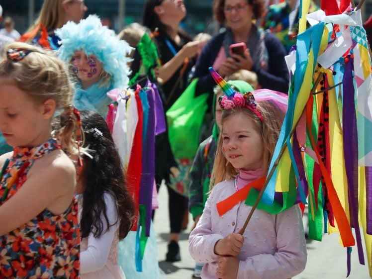 Image of Children in colourful costumes and face paint hold vibrant, ribbon-decorated sticks at an outdoor event or parade. Adults and other children, some with costumes and accessories, stand and watch in the background.