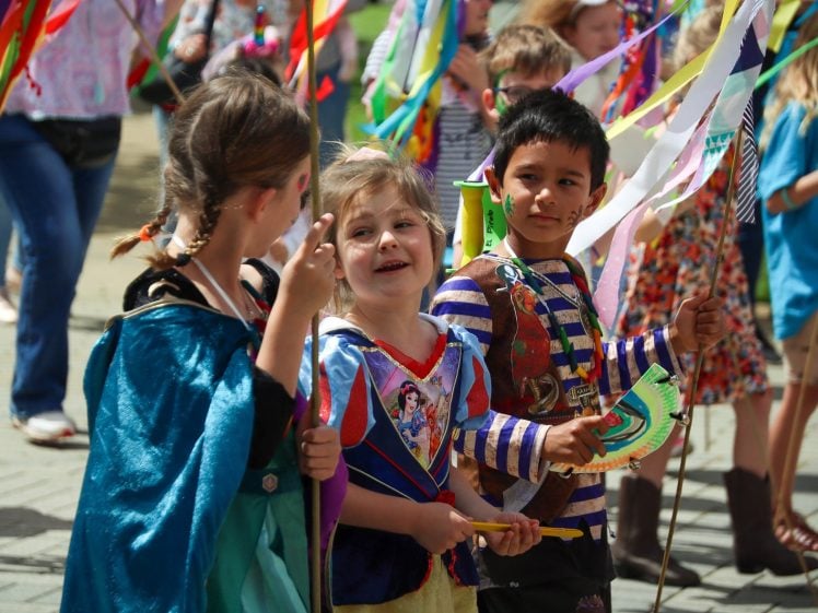 Image of Children in colourful costumes and holding flags walk together in a parade. They are smiling and appear to be enjoying the festive event, with other participants and bright decorations in the background.