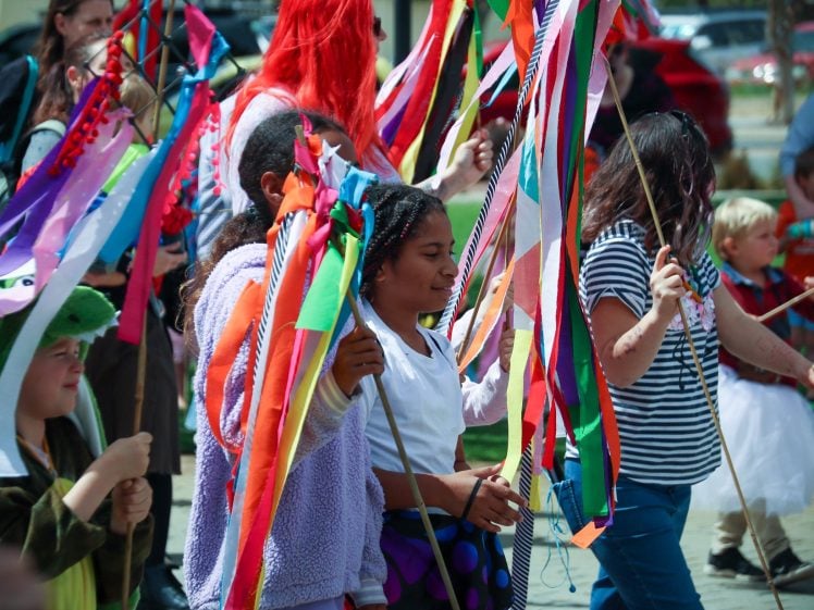 Image of Children and adults participate in a festive outdoor parade, holding colourful ribbon sticks. Some are dressed in fancy dress, and the atmosphere appears lively and joyful with people in the background.