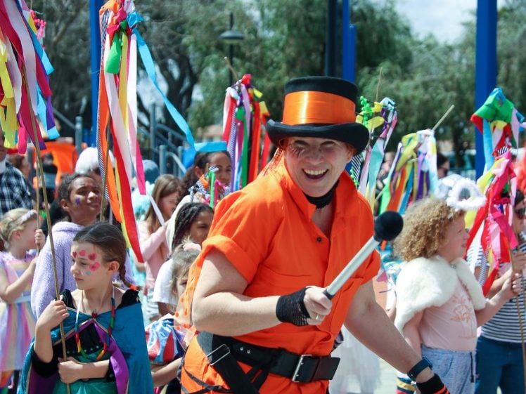 Image of A person in an orange outfit and black top hat leads a festive procession of children holding colourful ribbon sticks. The children wear bright costumes and face paint, celebrating outdoors on a sunny day.