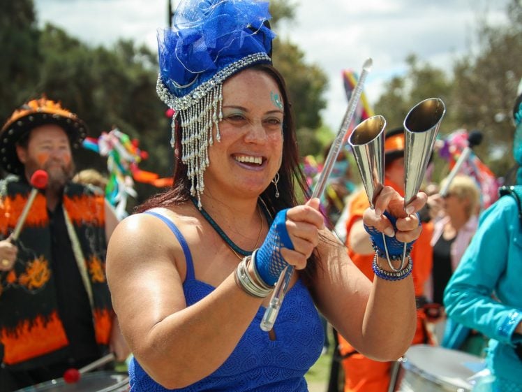 Image of A smiling woman in a bright blue dress and elaborate headpiece plays metal percussion instruments at an outdoor festival, surrounded by other musicians in colourful costumes.