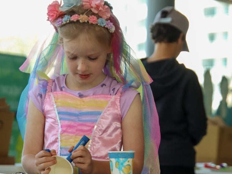 Image of A young girl in a pastel rainbow dress and flower headband concentrates on decorating a paper cup with a felt-tip pen. A boy in a cap stands blurred in the background.