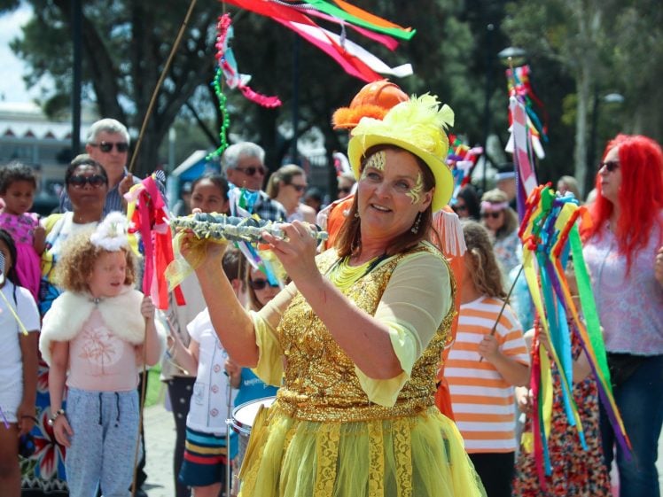 Image of A woman in a bright yellow costume and hat plays a musical instrument at an outdoor festival, surrounded by people holding colourful ribbons and wearing festive outfits. Trees and other festival-goers are visible in the background.