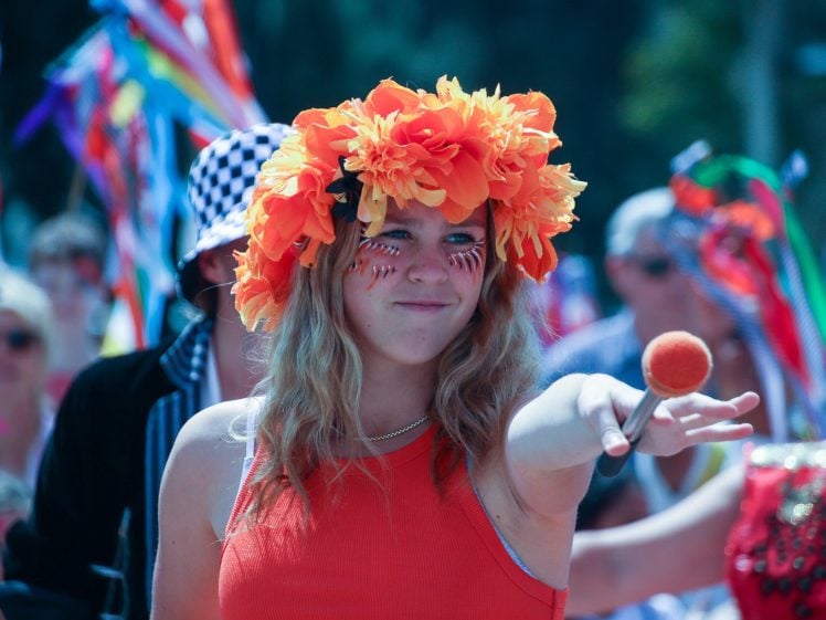 Image of A woman wearing a bright orange flower crown and orange top twirls a baton with an orange tip in a colourful, festive crowd. Her face is painted, and vibrant flags and costumes fill the background.