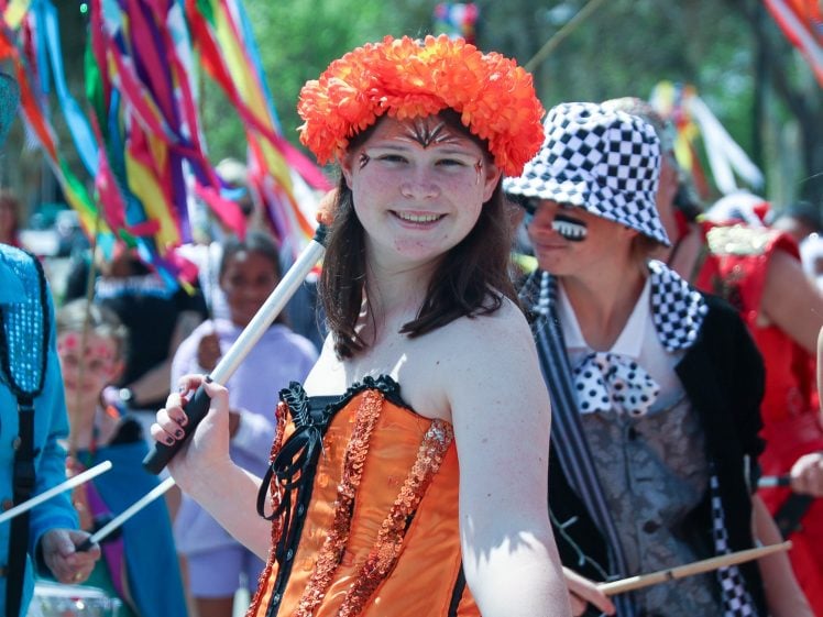 Image of A woman in an orange corset and flower crown smiles whilst holding a drumstick in a lively outdoor parade, surrounded by people in colourful costumes and ribbons.