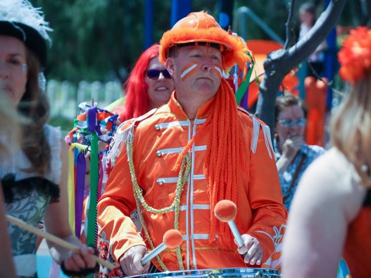 Image of A person dressed in a bright orange, marching band-style costume with orange face paint and decorative ropes plays a drum in a festive outdoor parade, surrounded by other colourfully dressed people.