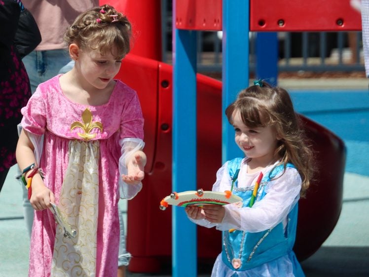 Image of Two young girls in princess costumes are playing together outdoors; one girl in a pink dress looks on whilst the other, in a blue dress, holds a toy crown. A playground structure is visible in the background.