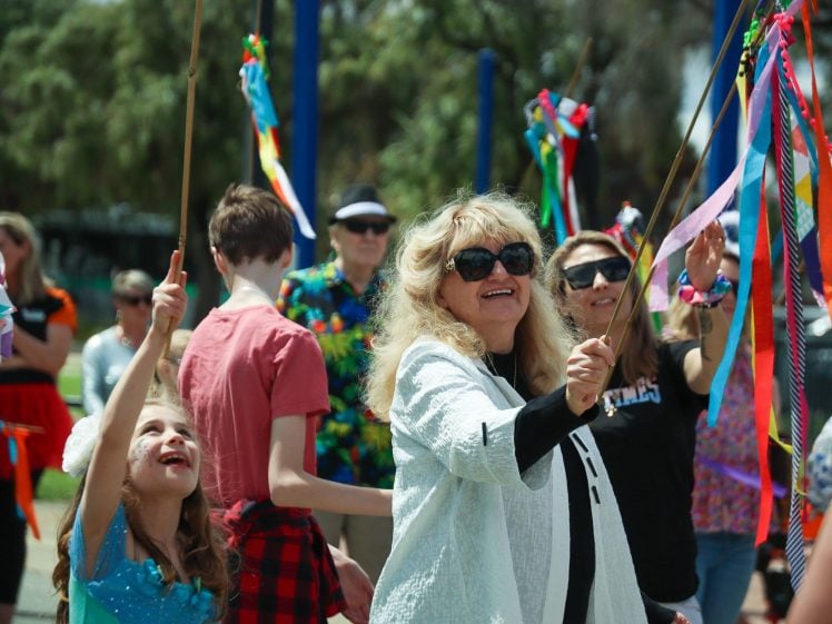 Image of A group of people outdoors joyfully wave colourful ribbon wands. A woman in sunglasses and a smiling young girl are in the foreground, surrounded by others, enjoying a festive, sunny day. Trees and decorations are visible in the background.