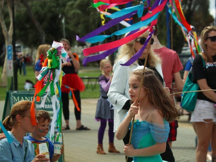 Image of A young girl in a blue dress holds a colourful ribbon wand at an outdoor event, surrounded by adults and children, some also carrying vibrant ribbon wands, with trees and people in the background.