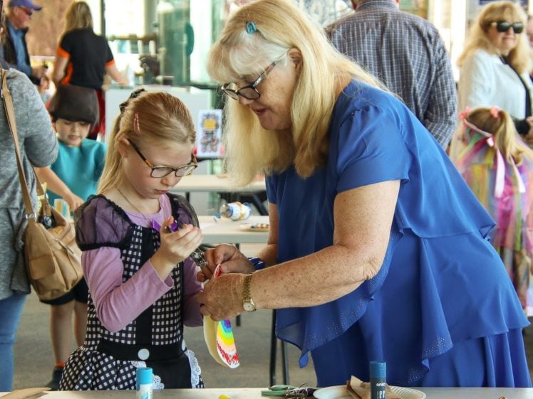 Image of An older woman in a blue dress helps a young girl decorate a colourful item at a craft table during a busy indoor event. Other people and children work on art projects in the background.